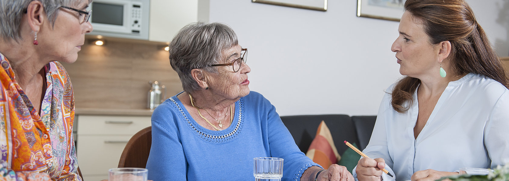 Die Pflegebedürftige, eine Angehörige und die Gutachterin des Medizinischen Dienstes sitzen während der Pflegebegutachtung an einem Tisch und sprechen miteinander.
